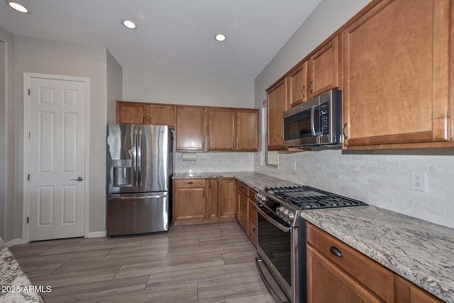 kitchen with tasteful backsplash, brown cabinetry, light wood-style flooring, appliances with stainless steel finishes, and light stone countertops