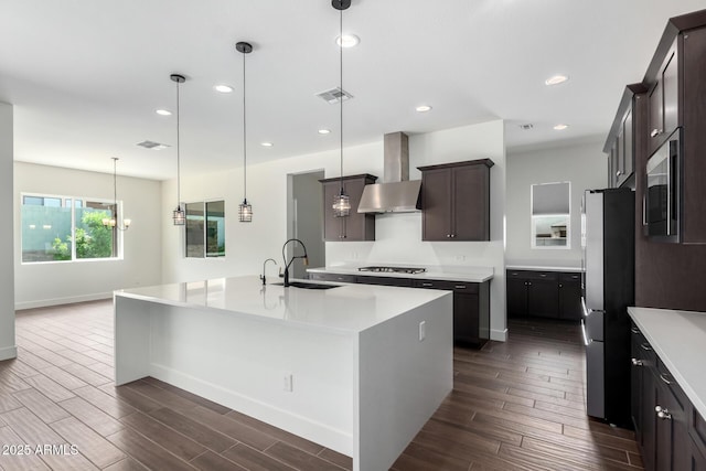 kitchen with wall chimney range hood, a sink, an island with sink, stainless steel appliances, and dark wood-style floors
