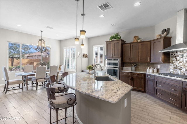 kitchen featuring sink, appliances with stainless steel finishes, a kitchen island with sink, decorative backsplash, and wall chimney range hood