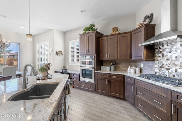 kitchen with pendant lighting, sink, appliances with stainless steel finishes, light stone countertops, and wall chimney exhaust hood