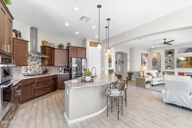 kitchen with stainless steel appliances, hanging light fixtures, a center island with sink, and wall chimney range hood