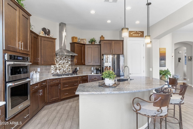 kitchen with a kitchen island with sink, light stone counters, wall chimney exhaust hood, and appliances with stainless steel finishes