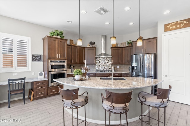 kitchen with pendant lighting, wall chimney range hood, sink, appliances with stainless steel finishes, and a center island with sink