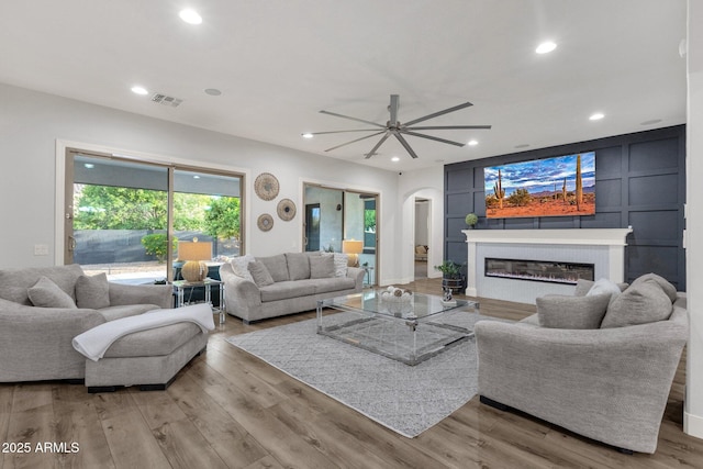 living room with arched walkways, a ceiling fan, a glass covered fireplace, and light wood-style floors