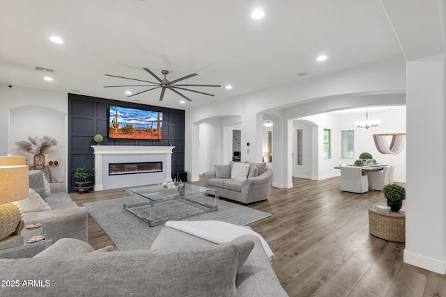 living room featuring arched walkways, a glass covered fireplace, ceiling fan with notable chandelier, wood finished floors, and visible vents