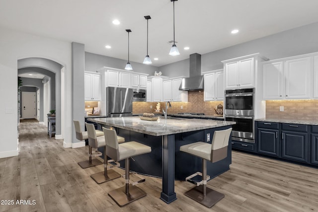 kitchen featuring a breakfast bar area, wall chimney exhaust hood, white cabinetry, light stone counters, and stainless steel appliances