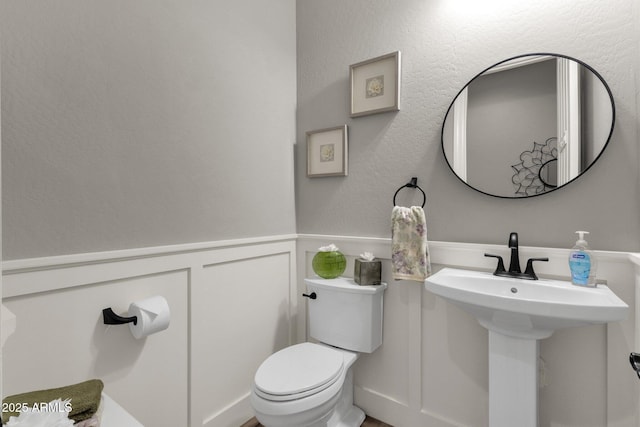 half bathroom featuring a decorative wall, toilet, a sink, and a wainscoted wall