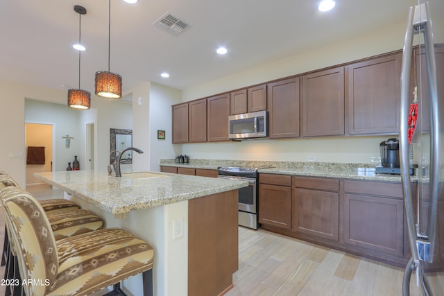 kitchen with a breakfast bar area, hanging light fixtures, appliances with stainless steel finishes, an island with sink, and light stone countertops