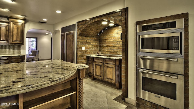 kitchen featuring dark brown cabinets, light stone countertops, backsplash, and light tile patterned floors