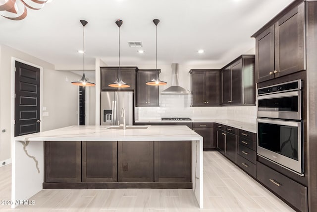 kitchen featuring wall chimney exhaust hood, sink, an island with sink, pendant lighting, and stainless steel appliances