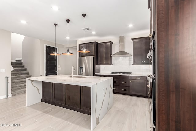 kitchen featuring an island with sink, sink, hanging light fixtures, stainless steel refrigerator with ice dispenser, and wall chimney range hood