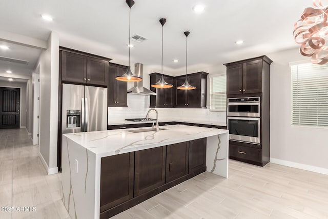 kitchen featuring sink, hanging light fixtures, wall chimney range hood, stainless steel appliances, and a center island with sink