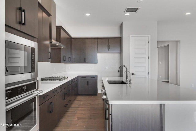 kitchen featuring wall chimney range hood, dark brown cabinets, appliances with stainless steel finishes, and a sink