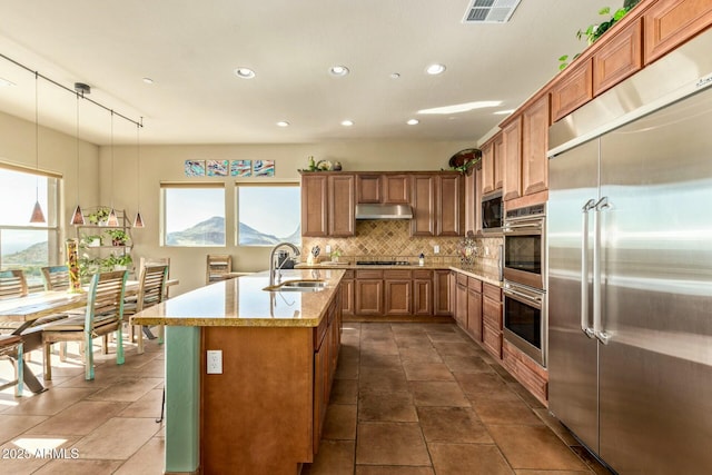 kitchen with sink, tasteful backsplash, built in appliances, hanging light fixtures, and a center island with sink