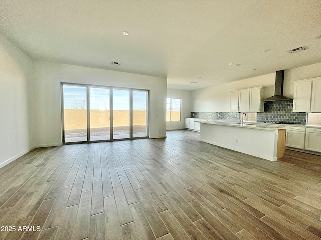 kitchen with white cabinetry, backsplash, wall chimney range hood, a center island with sink, and light wood-type flooring