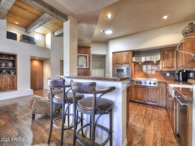 kitchen with wall chimney range hood, dark wood-type flooring, a breakfast bar, stainless steel appliances, and tasteful backsplash