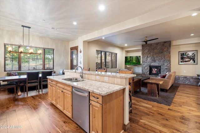 kitchen with sink, a stone fireplace, stainless steel dishwasher, a center island with sink, and light wood-type flooring