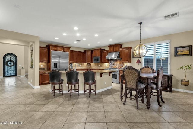 kitchen with a kitchen breakfast bar, ventilation hood, built in appliances, a center island with sink, and hanging light fixtures