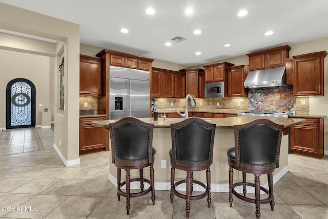 kitchen featuring a breakfast bar area, built in appliances, a kitchen island with sink, and ventilation hood