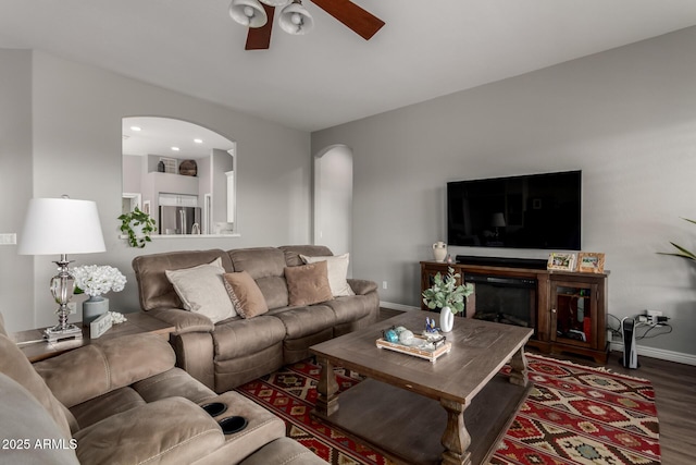 living area featuring arched walkways, a ceiling fan, a glass covered fireplace, wood finished floors, and baseboards