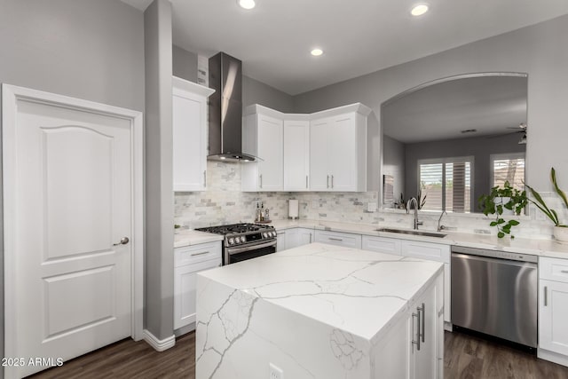 kitchen featuring arched walkways, appliances with stainless steel finishes, a sink, a kitchen island, and wall chimney exhaust hood