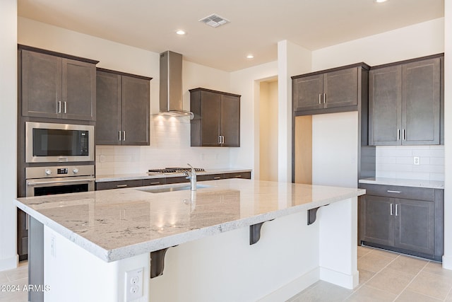 kitchen with built in microwave, sink, a kitchen island with sink, light stone countertops, and wall chimney range hood