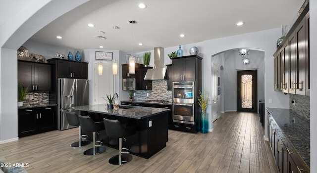 kitchen with pendant lighting, stainless steel appliances, a kitchen breakfast bar, a center island with sink, and wall chimney exhaust hood