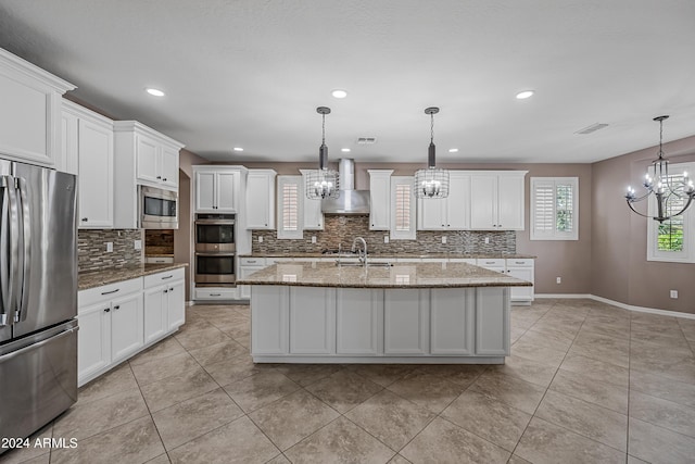 kitchen with wall chimney range hood, stainless steel appliances, hanging light fixtures, and an island with sink