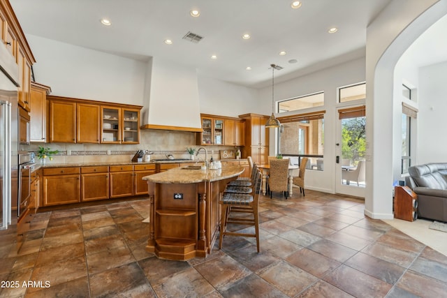 kitchen with visible vents, brown cabinets, light stone countertops, a high ceiling, and custom exhaust hood