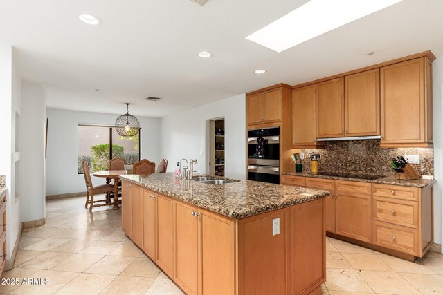 kitchen featuring a skylight, double oven, a kitchen island with sink, decorative light fixtures, and stone countertops