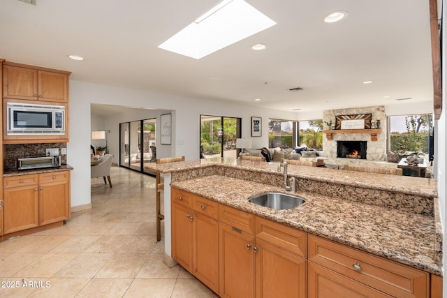 kitchen featuring a skylight, light stone countertops, stainless steel microwave, sink, and a stone fireplace