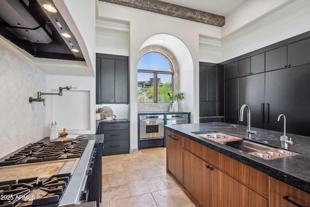 kitchen with dark stone countertops, decorative backsplash, a sink, appliances with stainless steel finishes, and exhaust hood