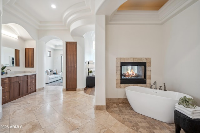 bathroom featuring a tiled fireplace, vanity, a tray ceiling, crown molding, and a freestanding bath