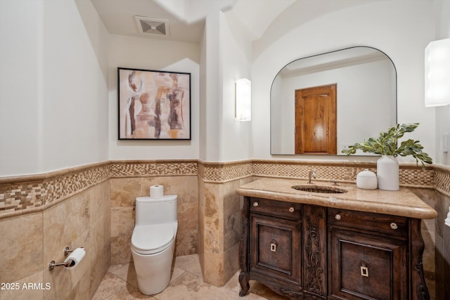 half bathroom with wainscoting, tile walls, toilet, vanity, and visible vents
