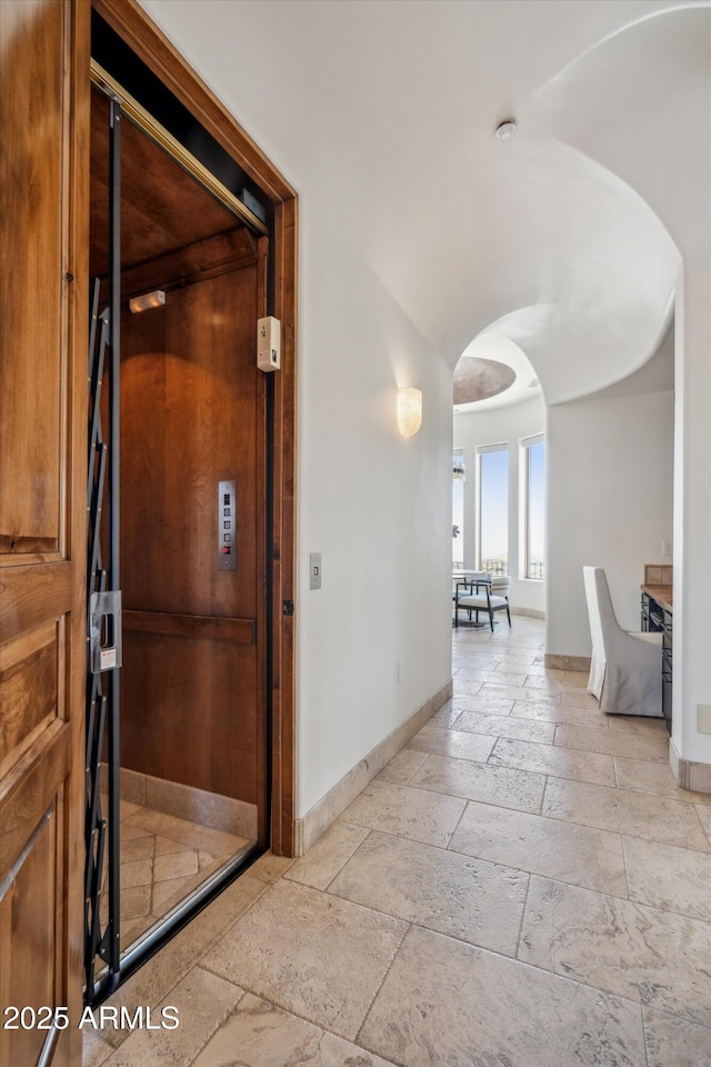 corridor with stone tile floors, baseboards, arched walkways, and elevator