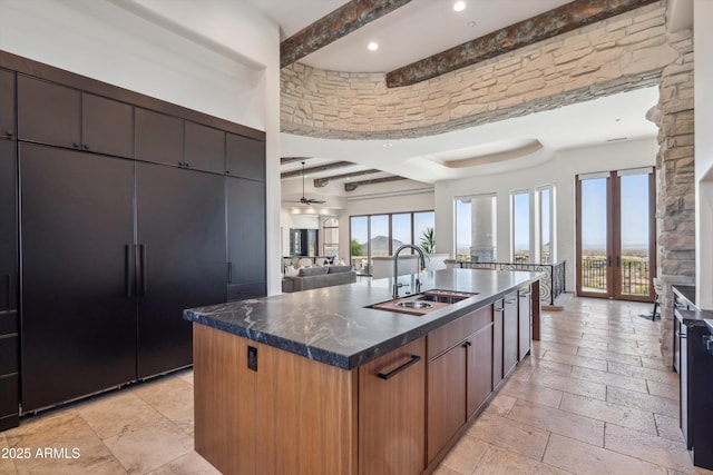 kitchen with a sink, stone tile flooring, beam ceiling, open floor plan, and a kitchen island with sink