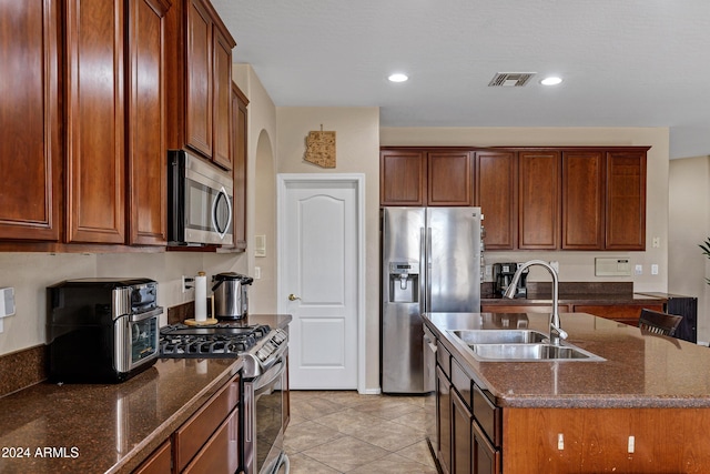 kitchen featuring a kitchen island with sink, sink, dark stone countertops, light tile patterned floors, and appliances with stainless steel finishes