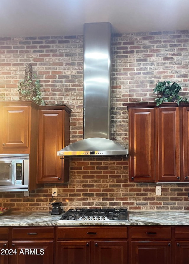 kitchen featuring light stone counters, wall chimney exhaust hood, stainless steel appliances, and brick wall