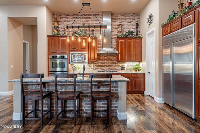 kitchen featuring an island with sink, light stone countertops, wall chimney exhaust hood, built in appliances, and a kitchen bar