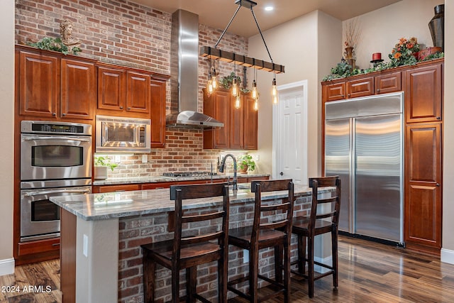 kitchen with light stone countertops, dark hardwood / wood-style flooring, wall chimney range hood, built in appliances, and a kitchen bar