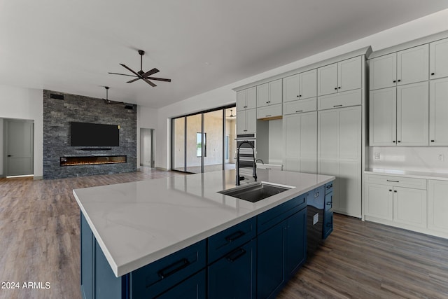 kitchen with dark hardwood / wood-style floors, ceiling fan, a stone fireplace, an island with sink, and sink