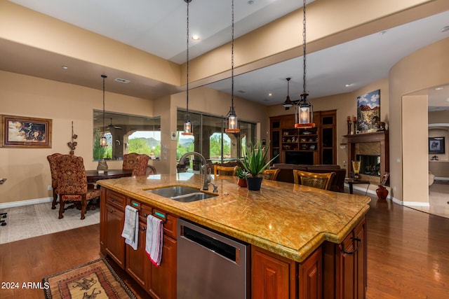 kitchen featuring a center island with sink, a sink, stainless steel dishwasher, a fireplace, and dark wood-style flooring