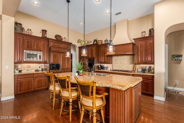 kitchen with a sink, built in appliances, custom exhaust hood, arched walkways, and a kitchen island with sink