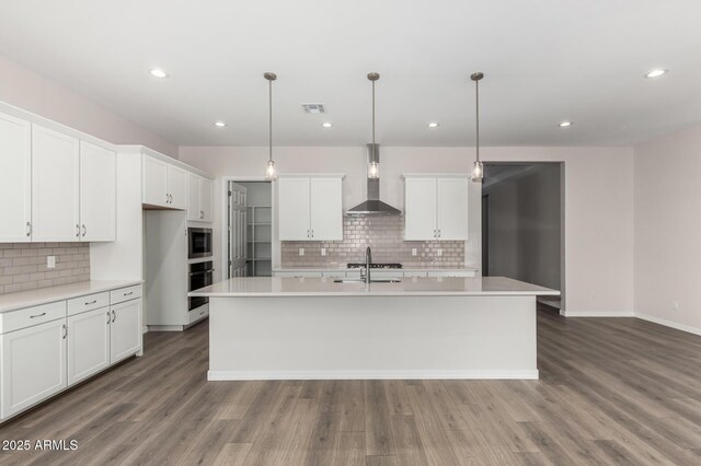 kitchen featuring a center island with sink, wall chimney exhaust hood, stainless steel microwave, white cabinetry, and decorative light fixtures