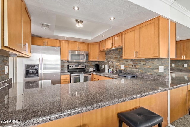 kitchen with a sink, a tray ceiling, stainless steel appliances, dark stone counters, and a peninsula