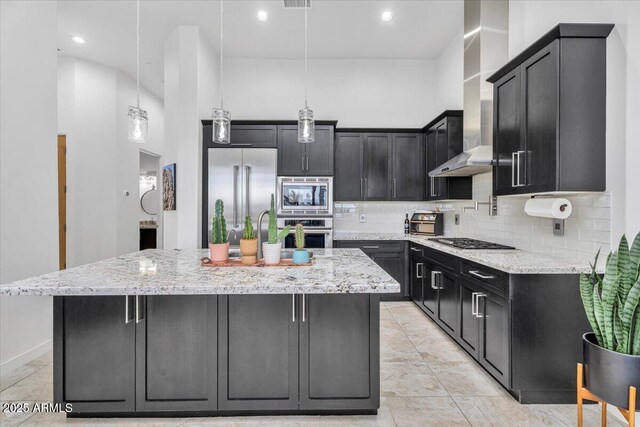 kitchen with built in appliances, light stone counters, a high ceiling, wall chimney exhaust hood, and a kitchen island with sink