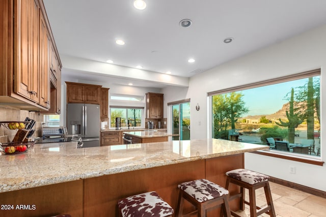 kitchen with light stone countertops, decorative backsplash, kitchen peninsula, stainless steel fridge with ice dispenser, and a kitchen bar