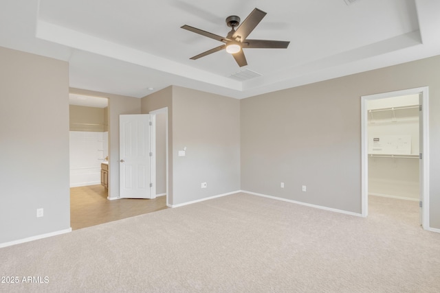 unfurnished bedroom featuring ensuite bath, ceiling fan, a tray ceiling, light carpet, and a walk in closet