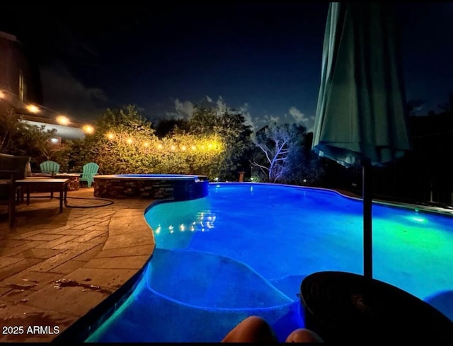 pool at night featuring a pool with connected hot tub and a patio