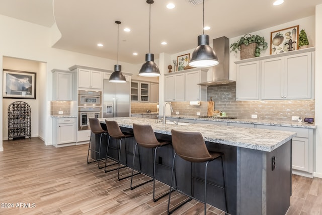 kitchen featuring stainless steel appliances, wall chimney range hood, pendant lighting, white cabinets, and an island with sink